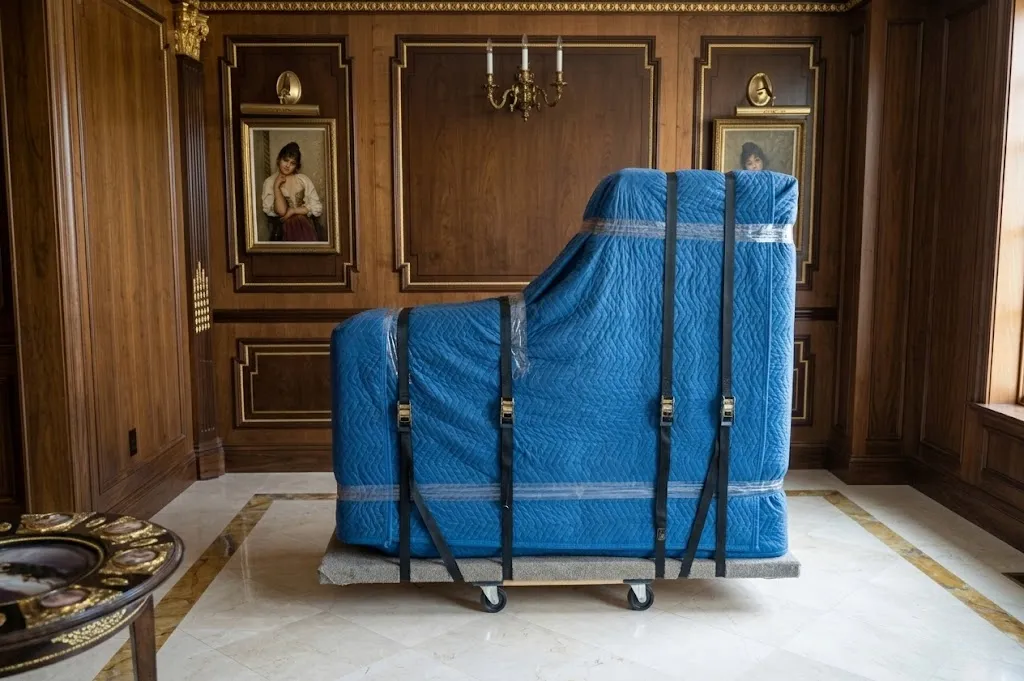 Grand piano protected by blue quilted padding and multiple black tension straps, resting on a heavy-duty dolly.