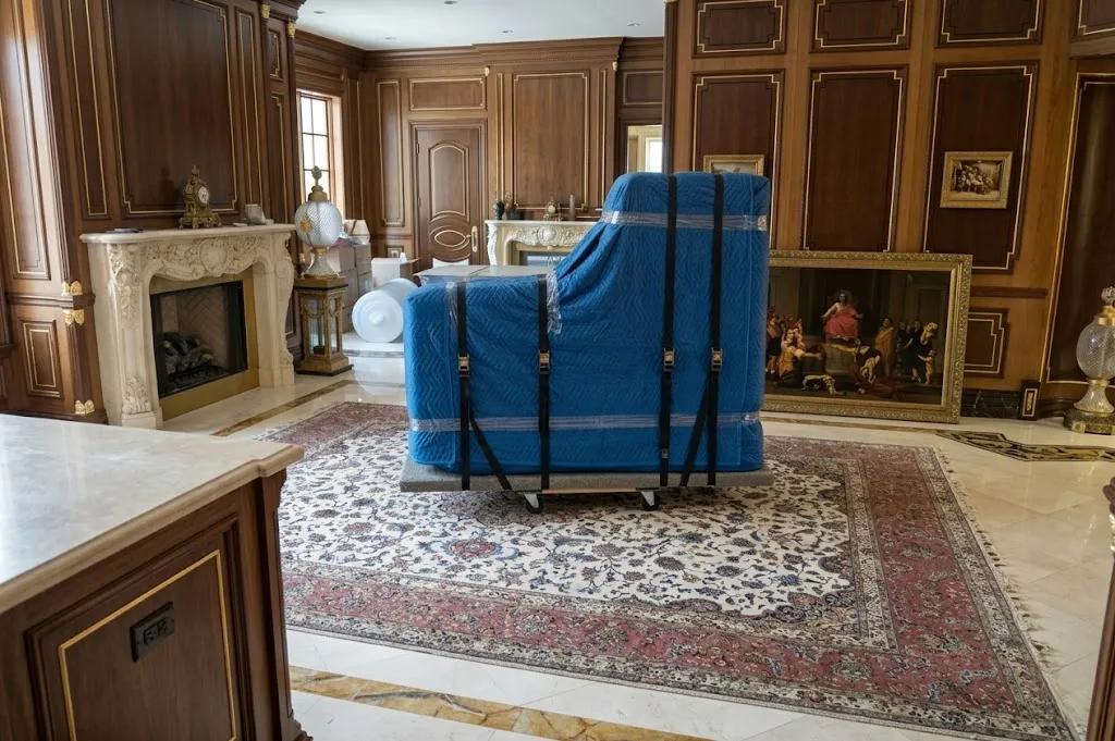 Grand piano wrapped in blue quilted blankets and secured with straps, positioned on a rolling dolly.